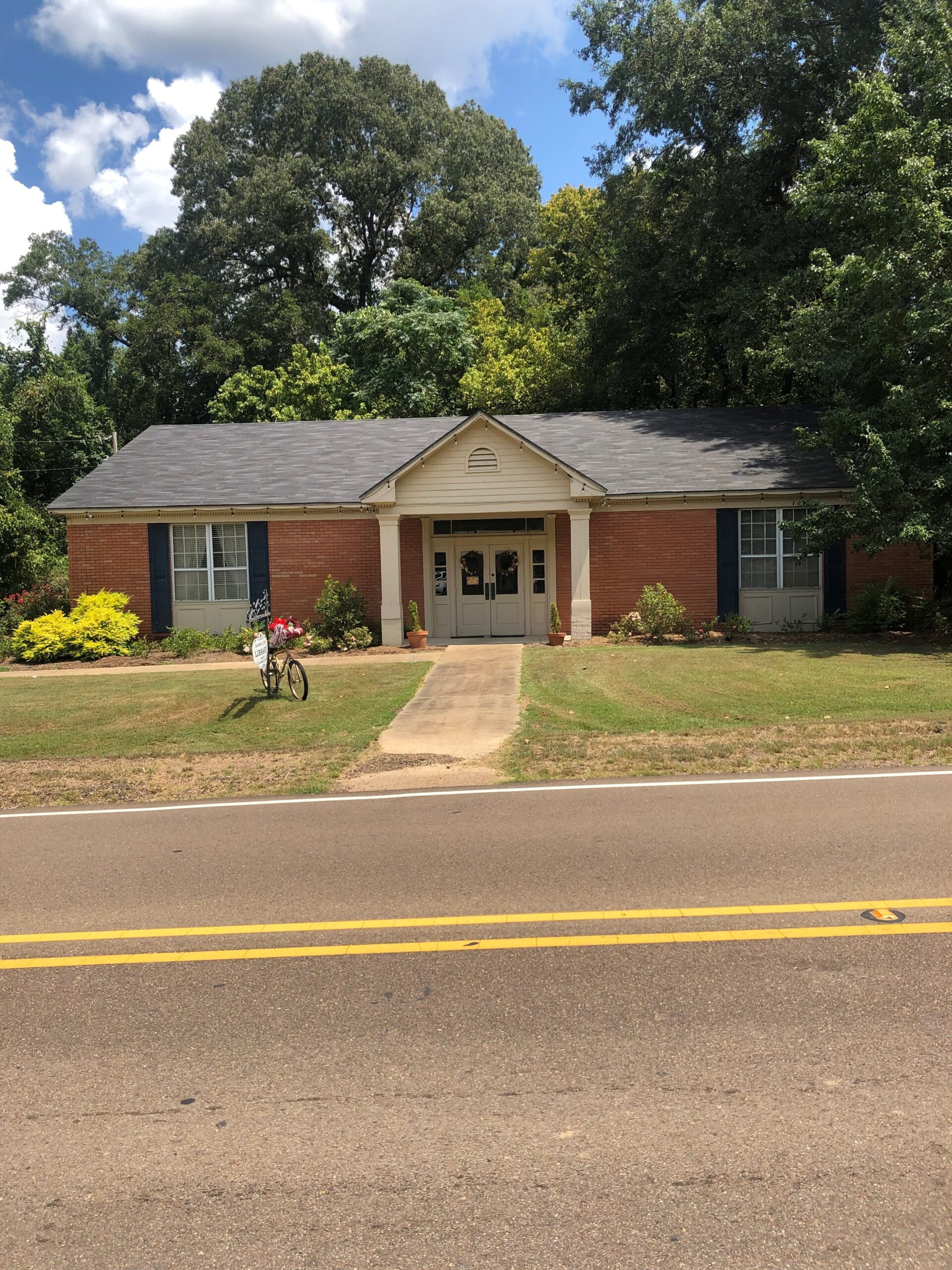 Red brick, single-story library with columned entrance, a bicycle in the front, and trees in the background.