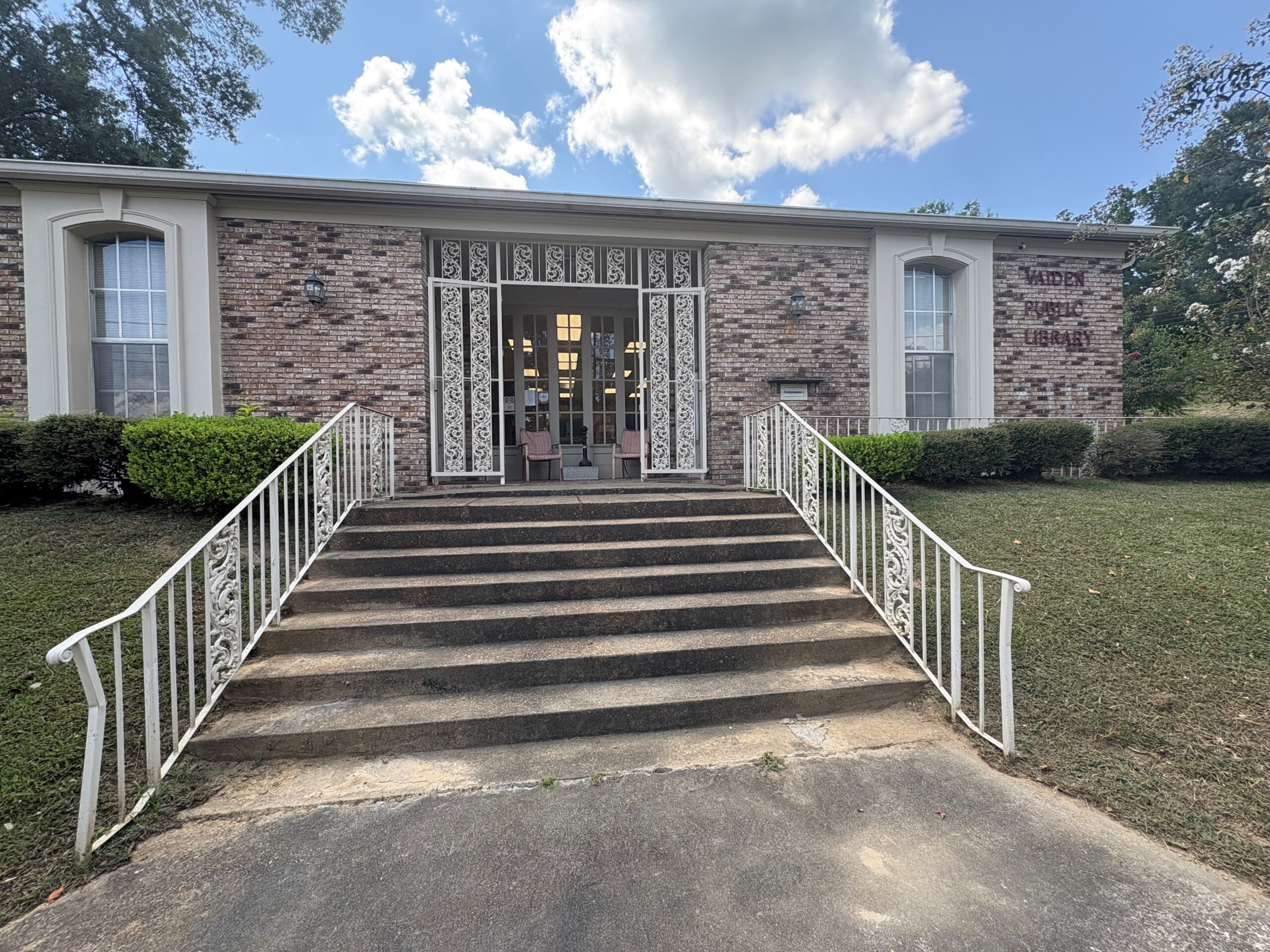 Brick library with white trim, front steps, and decorative railings under a partly cloudy sky.